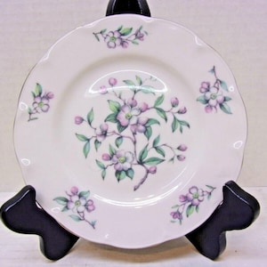 May include: A white ceramic plate with a scalloped edge, decorated with a floral pattern of pink and purple blossoms and green leaves. The plate is displayed on a black stand, showcasing its delicate design.