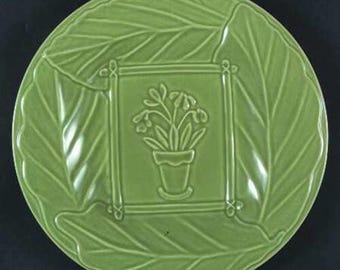 May include: A round, light green plate with a leaf-like design around the rim. The center features a square frame with a potted flower design. The plate is made of ceramic and has a glossy finish.
