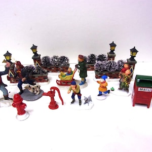 May include: A collection of miniature Christmas village figurines depicting various winter scenes. The set includes figures of people, dogs, a fire hydrant, a water pump, a sled, and a mailbox. The figurines are painted with bright colors and are set against a backdrop of brick walls and lampposts.