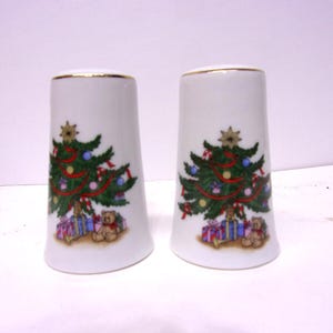 May include: Two white ceramic salt and pepper shakers with a gold rim. Each shaker features a Christmas tree design with ornaments, presents, and a teddy bear. The Christmas tree is decorated with red ribbon, blue and gold ornaments.