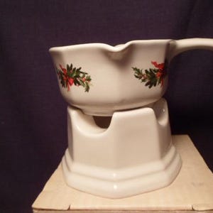 May include: White ceramic gravy boat with a handle, resting on a matching stand. The boat features a festive design of red berries and green leaves. The stand has a hexagonal base.