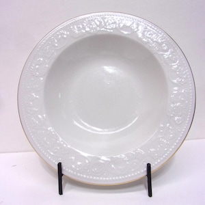 May include: A white ceramic bowl with a decorative embossed floral pattern around the rim. The bowl has a gold trim and is displayed on a black metal stand. The bowl is likely for serving food.