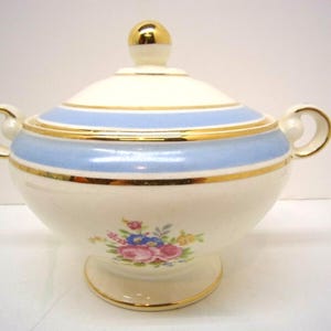 May include: A vintage lidded sugar bowl with a cream-colored base, adorned with a light blue band and gold trim. The bowl features a floral design and a gold knob on the lid. It has two handles and a pedestal base.