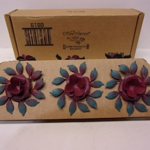 May include: A decorative floral accent piece featuring three burgundy rose blossoms with teal and burgundy leaves. The piece is mounted on a brown cardboard base. A brown cardboard box is in the background.
