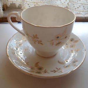 May include: White teacup and saucer set with gold trim and floral accents. The cup and saucer feature a delicate design of gold leaves and small blue flower details. A classic and elegant tea set.