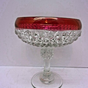 May include: A clear glass dessert dish with a ruby red rim. The bowl has a diamond-cut pattern and sits atop a clear stem and circular base. The dish is designed for serving desserts.