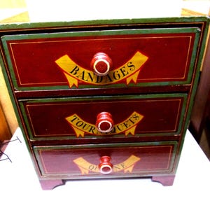 May include: A three-drawer wooden cabinet with a burgundy finish and green trim. Each drawer features a yellow banner with text and a red knob. The top drawer reads "BANDAGES". The middle drawer reads "TOURNIQUETS". The bottom drawer reads "QUINE".