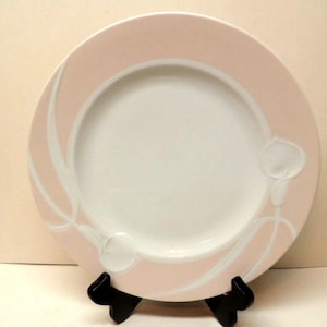 May include: A round, white plate with a pink border and white floral design. The plate is displayed on a black stand. The plate's design features a white floral pattern and a white center.