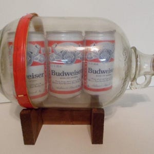 May include: A vintage glass beer bottle with a red handle, containing three Budweiser beer cans. The bottle rests on a small wooden stand. The cans have red and white labels.