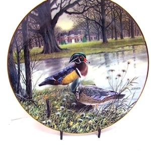 May include: Decorative plate featuring a detailed painting of two wood ducks near a pond. The plate has a gold rim and a serene landscape background with trees and a house. The artist's signature "JERNER" is visible.