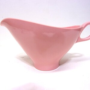 May include: A vintage pink ceramic gravy boat with a handle. The gravy boat has a curved spout and a rounded handle. The color is a soft, solid pink. The gravy boat is likely from the mid-20th century.