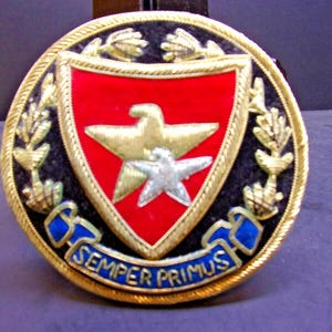 May include: A circular emblem with a red shield featuring a gold eagle and stars, surrounded by gold laurel leaves and a blue banner with the text "SEMPER PRIMUS". The emblem has a black background and gold trim.