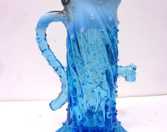 May include: A handcrafted, blue glass pitcher with a unique, textured design. The pitcher features a handle and a spout, with a flared, wavy rim. The glass has a bubbly texture and a gradient of light to dark blue.