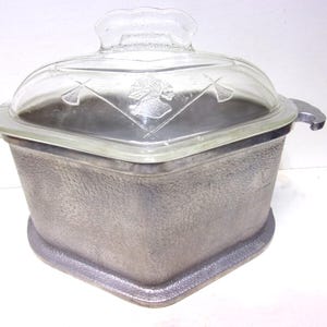 May include: A square, gray metal casserole dish with a clear glass lid. The lid has a handle and a decorative design. The dish has two handles and sits on a slightly raised base. The dish is designed for cooking.
