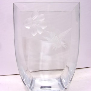 May include: Clear glass vase with a square base and gently curved sides. Etched on the front are a flower and a hummingbird. The vase has a label at the base that reads "MIKASA".