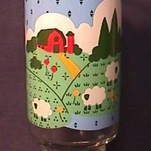 May include: Clear glass tumbler with a farm scene design. The illustration features a red barn, green hills, white sheep, and a blue sky with clouds. The glass has a wide base and a straight, cylindrical top.