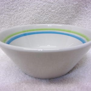 May include: White ceramic bowl with a simple design. The bowl features two horizontal stripes in blue and green near the rim. The bowl is set against a white background.