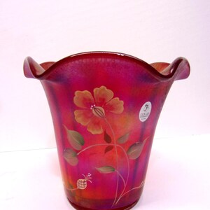 May include: A vibrant, hand-painted glass vase with a flared shape and ruffled rim. The vase features a floral design in shades of orange, pink, and green. The background transitions from deep red to lighter pink hues.