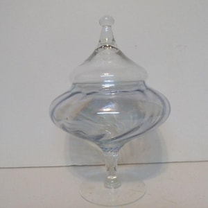 May include: A clear glass apothecary jar with a lid. The jar has a bulbous body and a slender stem, resting on a circular base. The lid is topped with a small, rounded knob. The glass has a slight blue tint.