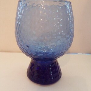 May include: A blue glass goblet with a textured surface. The goblet has a rounded bowl and a short, sturdy base. The glass appears to be a light blue color, with a darker blue base.