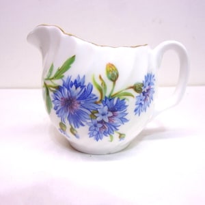May include: White porcelain creamer with a delicate blue floral design featuring cornflowers and green leaves. The creamer has a small handle and a spout, with a gold trim around the rim. A charming addition to any tea set.