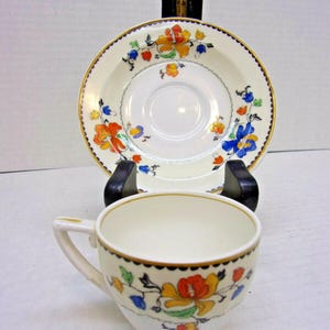 May include: A vintage teacup and saucer set. The off-white ceramic cup and saucer feature a floral design with orange, yellow, blue, and green accents. The rims are trimmed with gold and a dark brown scalloped border. The set is displayed on a black stand.