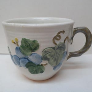 May include: A white ceramic teacup with a painted grape design. The grapes are blue and the leaves are green. The handle is a dark brown color. The cup is approximately 4 inches wide and 3 inches tall.