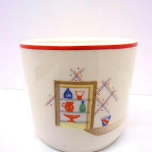 May include: Cream-colored ceramic cup with a red rim. The cup features a colorful illustration of a kitchen cabinet with various items. Blue, red, and yellow accents decorate the cup's exterior, adding a vintage aesthetic.