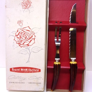 May include: Vintage cutlery set by Regent Sheffield, presented in a white box with red rose illustrations. The set includes a fork and two knives with serrated blades and brown and yellow handles. The box reads "Cutlery Set" and "Stainless Blades".