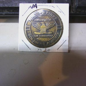May include: A gold-toned coin with an eagle emblem, presented by "The Commander." The coin is encased in a white paper holder with the handwritten letters "VA" at the top. The text on the coin reads "Mission First, Soldiers Always."