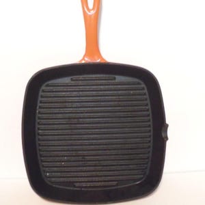 May include: A square, black cast iron grill pan with a ridged cooking surface. The handle is orange and has a hole for hanging. The pan is designed for grilling food and has a small lip on the side.