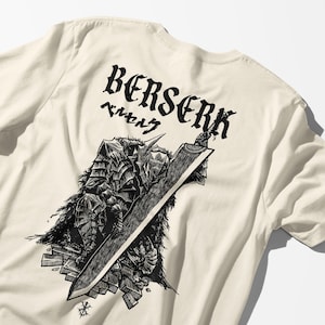 Puede incluir: Camiseta color crema con un gráfico negro de un guerrero con una espada grande. La palabra "BERSERK" está impresa encima de la ilustración, junto con caracteres japoneses. El diseño es detallado y estilizado.