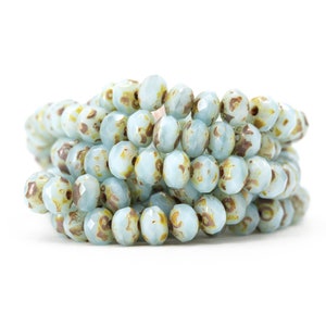 May include: A close-up of a pile of light blue glass beads with brown speckles. The beads are faceted and have a slightly iridescent finish.