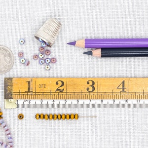 Pu&ograve; includere: Un righello di legno con i numeri da 1 a 5, una matita viola e nera, un ditale e una variet&agrave; di perline in tonalit&agrave; viola e oro.