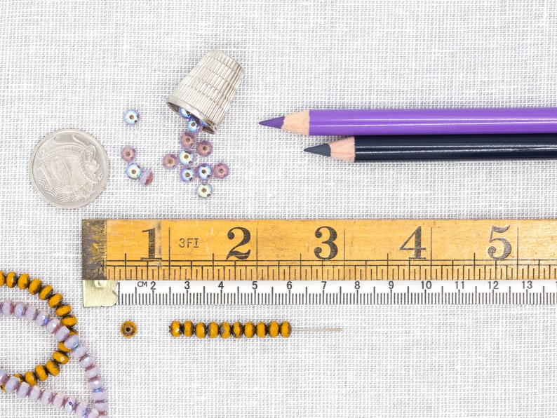 Pu&ograve; includere: Un righello di legno con i numeri da 1 a 5, una matita viola e nera, un ditale e una variet&agrave; di perline in tonalit&agrave; viola e oro.