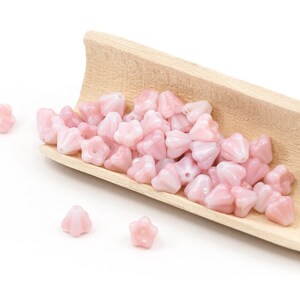 K&ouml;nnte beinhalten: Ein Holzsch&ouml;pfer gef&uuml;llt mit rosa gl&auml;sernen Blumenperlen. Die Perlen sind auf einer wei&szlig;en Oberfl&auml;che verstreut.