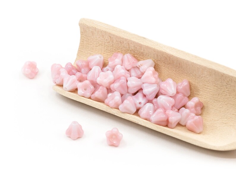 K&ouml;nnte beinhalten: Ein Holzsch&ouml;pfer gef&uuml;llt mit rosa gl&auml;sernen Blumenperlen. Die Perlen sind auf einer wei&szlig;en Oberfl&auml;che verstreut.