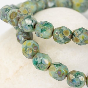 May include: Close-up of a strand of faceted beads in shades of green, blue, and yellow. The beads have a textured, mottled appearance and are strung together. The beads are resting on a white, rounded surface. Jewellery.