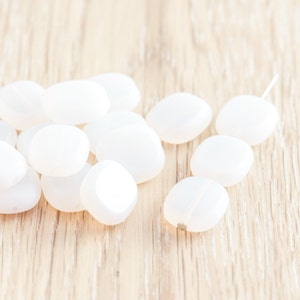 May include: A group of white oval-shaped beads on a light wood surface. The beads are smooth and glossy.