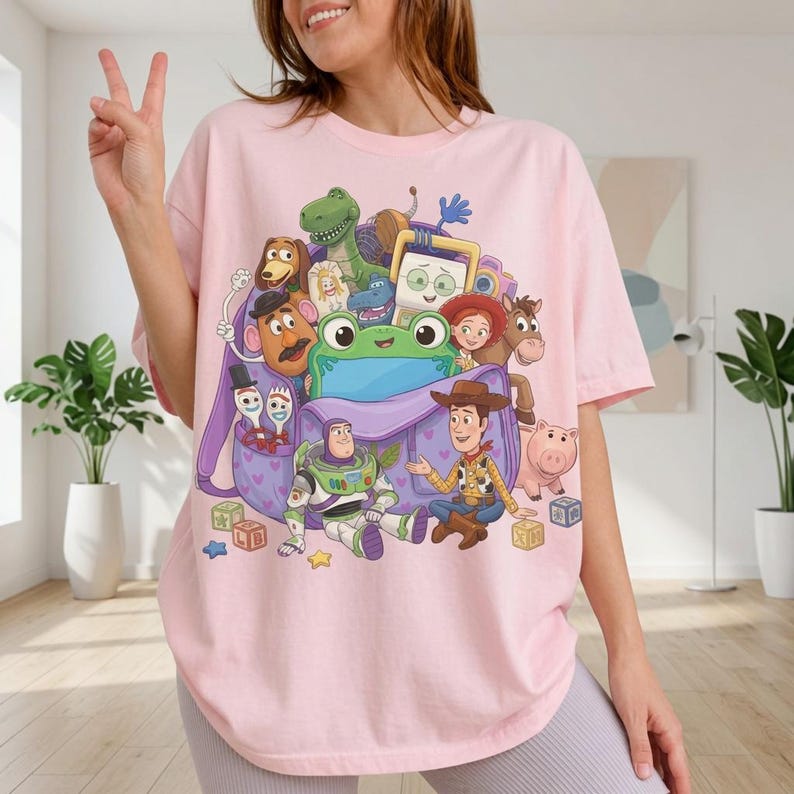 Puede incluir: Una camiseta rosa claro con un estampado gr&aacute;fico colorido de varios personajes de juguetes animados que salen de una mochila morada. El dise&ntilde;o incluye figuras como un vaquero, un guardi&aacute;n espacial, un dinosaurio verde, una patata y un cerdito.