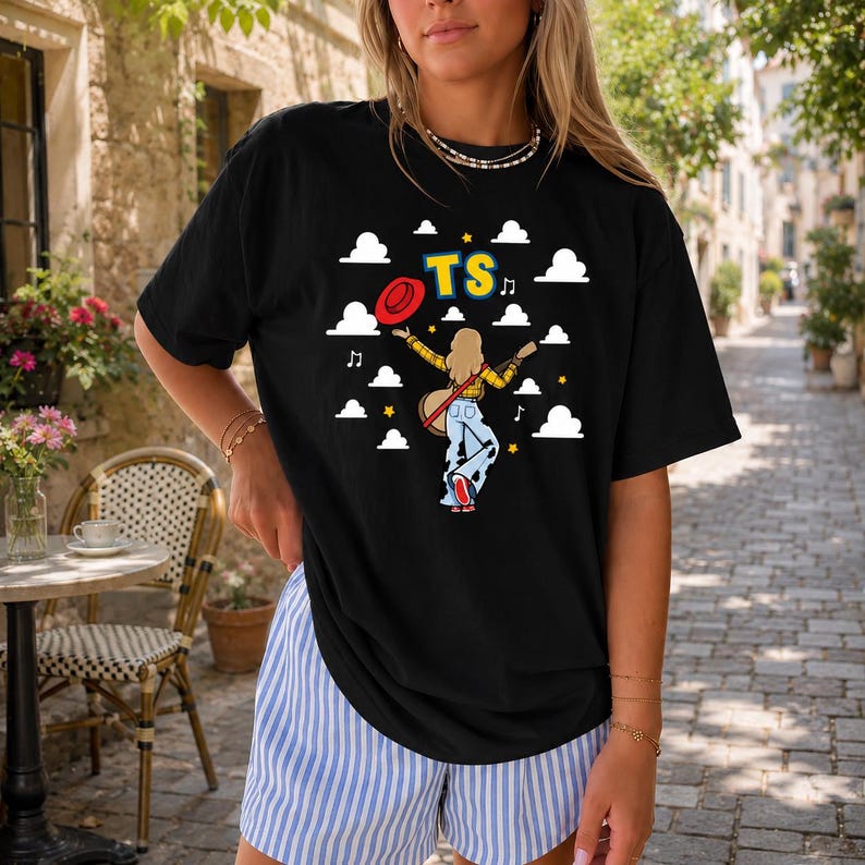 May include: A black short-sleeve t-shirt featuring a graphic of a person with blonde hair wearing a yellow plaid shirt and blue jeans, holding a red hat and guitar. The design includes white clouds, yellow stars, musical notes, and the text TS.