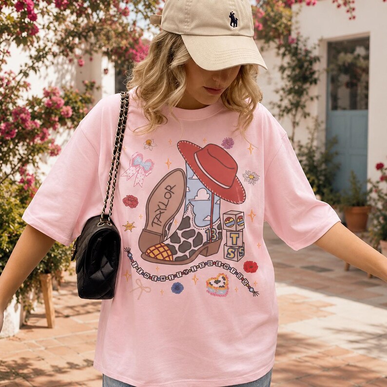 May include: A light pink short-sleeve t-shirt featuring a graphic print of a cow-print cowboy boot, a red cowboy hat, a string of beads, and blocks with the letters T, S, and the number 13. The word TAYLOR is printed on the boot.