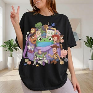 Puede incluir: Una camiseta negra con un estampado gr&aacute;fico de varios personajes animados de la franquicia Toy Story, incluidos Woody, Buzz Lightyear, Jessie, Rex y el Sr. Cara de Papa, todos reunidos alrededor y dentro de una mochila morada grande.