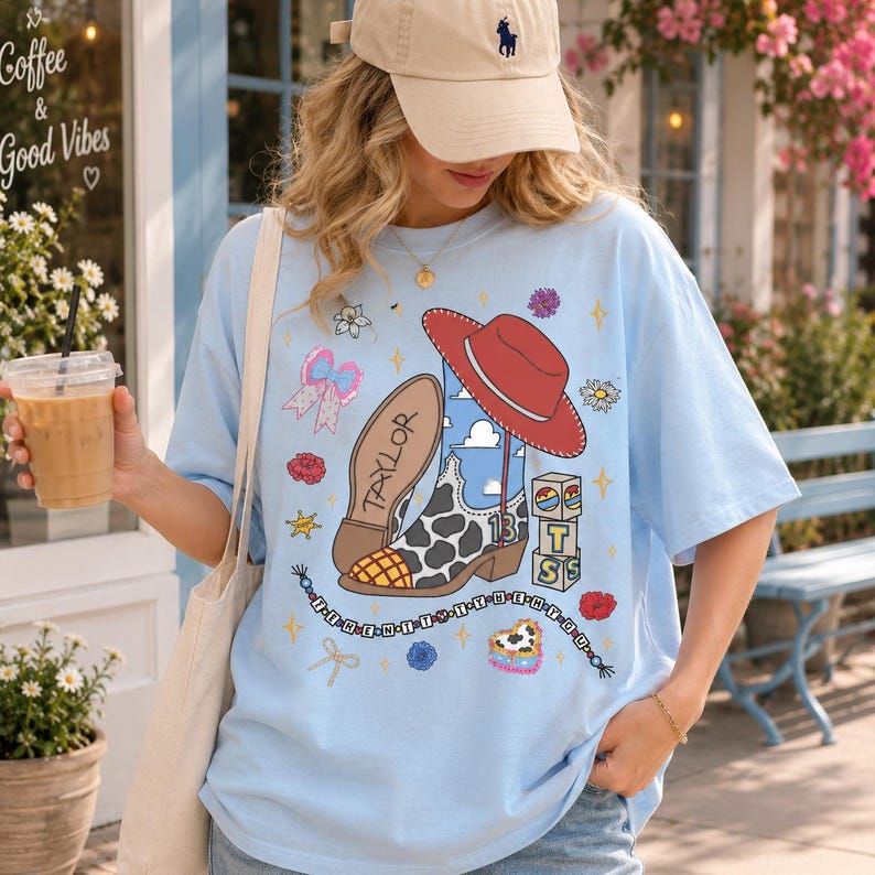 May include: A light blue t-shirt featuring a graphic print of a cowboy boot with a cow print pattern, a red hat, building blocks, a bow, and the name Taylor. The design includes a beaded string with text and various small floral and star icons.
