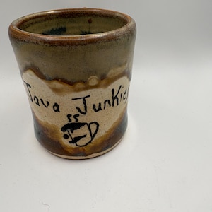 May include: A handmade ceramic mug with a brown and tan glaze. The mug features the words "Java Junkie" in black script, along with a coffee cup illustration. The mug has a slightly tapered shape and a rustic aesthetic.