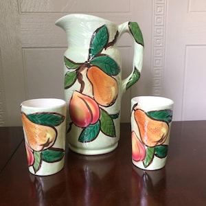 Könnte beinhalten: Eine hellgrüne Keramikkaraffe und zwei passende Becher. Die Karaffe hat einen großen Griff und ist mit Bildern von Birnen und Pfirsichen mit grünen Blättern verziert. Die Becher haben das gleiche Fruchtdesign.