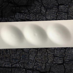 May include: A rectangular, off-white ceramic serving dish with three curved indentations. The dish has a ribbed texture and is set against a dark, textured background, suggesting a rustic or natural aesthetic. Ideal for serving appetizers or small portions.