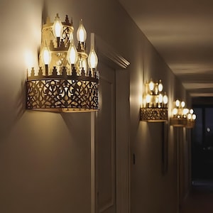 Puede incluir: Apliques dorados ornamentados con múltiples bombillas en forma de vela a lo largo de un pasillo. Los apliques presentan un diseño calado decorativo y están montados en una pared de color neutro. La luz cálida crea un ambiente acogedor.