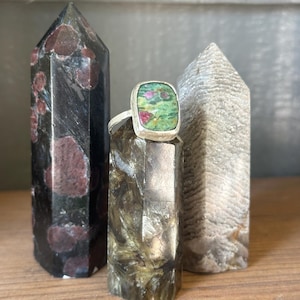 May include: A silver ring with a square, green and red stone is displayed between three crystal towers. The ring's stone has a unique pattern. The crystal towers are black, brown, and gray. The ring and crystals are on a wooden surface.
