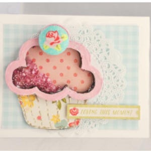 May include: A pink and white cupcake with a pink and white polka dot pattern and a blue and white flower on top. The cupcake is on a white background with a blue and white gingham pattern. The text "Loving this moment" is on a yellow banner.